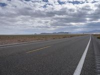 a road in a desert with mountains on both sides of it and clouds overhead and an area full of arid land below