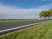 a road in the middle of green grass with trees and blue sky behind it near the roadway