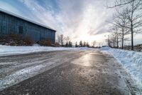 a snowy street is empty with many snow patches on it and a barn in the background