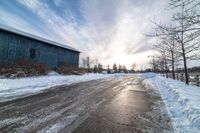 a snowy street is empty with many snow patches on it and a barn in the background