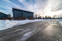 a road in front of some barn and buildings in the snow during the day time