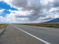 an empty highway running along the side of the road and into the distance, with mountains and grass, on a cloudy day