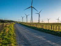 several windmills at sunset are in the distance from a winding country road with a fence