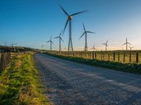 several windmills at sunset are in the distance from a winding country road with a fence