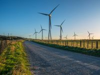 several windmills at sunset are in the distance from a winding country road with a fence