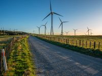 several windmills at sunset are in the distance from a winding country road with a fence