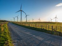 several windmills at sunset are in the distance from a winding country road with a fence