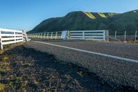 a fence and road near green hills in the distance with grass and dry grass on both sides