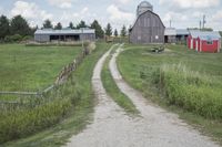 gravel road in front of two farm buildings next to tall grass and trees on the other side of fence and dirt trail