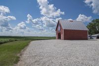 a barn on top of a gravel road next to grass and trees in front of a blue sky