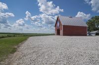 a barn on top of a gravel road next to grass and trees in front of a blue sky