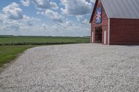 a barn on top of a gravel road next to grass and trees in front of a blue sky