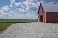 a barn on top of a gravel road next to grass and trees in front of a blue sky