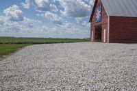 a barn on top of a gravel road next to grass and trees in front of a blue sky