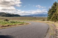 a lone empty asphalt road with lots of grass on either side of the path and the view of the ocean, mountains and trees,