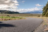 a lone empty asphalt road with lots of grass on either side of the path and the view of the ocean, mountains and trees,