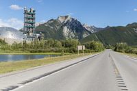 a long empty road with green trees, mountains and a building in the distance with blue sky above