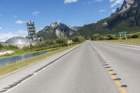 a long empty road with green trees, mountains and a building in the distance with blue sky above