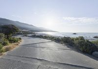 a street with gravel road and vegetation along the road near mountains by water area and mountain range