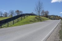 a road and fence running along side a hill near trees and grass on a sunny day