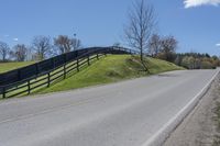 a road and fence running along side a hill near trees and grass on a sunny day