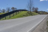 a road and fence running along side a hill near trees and grass on a sunny day