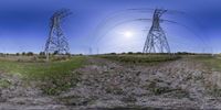 two high voltage poles are near grass and trees under a bright sky, with sun in the background