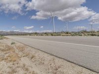 several wind turbines on an open road near a desert hillside under a cloudy blue sky