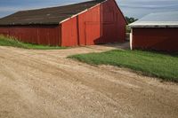 a red barn is near a dirt road with a white horse in front of it