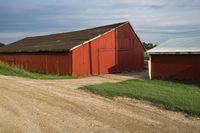 a red barn is near a dirt road with a white horse in front of it