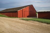 a red barn is near a dirt road with a white horse in front of it