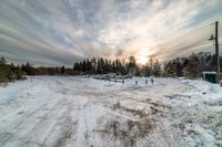 the sun shines brightly through a partly cloudy sky above a road and forest near snow covered buildings