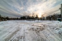 the sun shines brightly through a partly cloudy sky above a road and forest near snow covered buildings