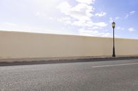 an empty street next to an empty wall and lamp post against the sky with clouds