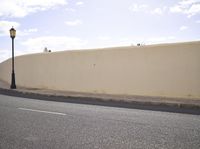 an empty street next to an empty wall and lamp post against the sky with clouds