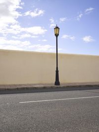 an empty street next to an empty wall and lamp post against the sky with clouds