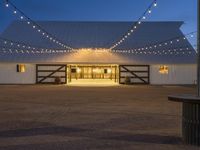 string lights hang across the front entrance to a barn at dusk with a wooden table and benches nearby