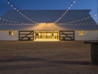 string lights hang across the front entrance to a barn at dusk with a wooden table and benches nearby