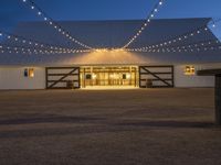 string lights hang across the front entrance to a barn at dusk with a wooden table and benches nearby