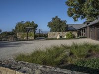 a photo taken of a rustic home in a country setting with a gravel driveway with trees to the side and a gate, stone pathway way, shrubs and stone walls