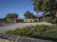 a photo taken of a rustic home in a country setting with a gravel driveway with trees to the side and a gate, stone pathway way, shrubs and stone walls