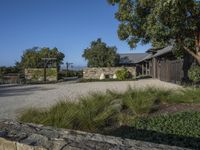 a photo taken of a rustic home in a country setting with a gravel driveway with trees to the side and a gate, stone pathway way, shrubs and stone walls