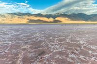 a vast desert with some sand blowing over it and clouds in the distance and mountains