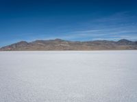 some large mountains and some snow and water on some flat ground with blue skies and one of them in the background