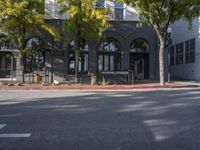 the buildings on a corner are tall and gray and green trees line the sidewalk and trees line the street