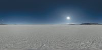 a sunlit over a large field of snow with hills in the background and a blue sky