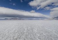 large expanse of flat salt under a cloudy blue sky and mountains and mountain peaks behind the salt