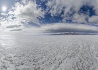 large expanse of flat salt under a cloudy blue sky and mountains and mountain peaks behind the salt