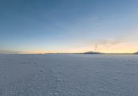 a lone man walking across a large, frozen field at dusk, with the sun low in the sky and some clouds above