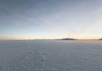 a lone man walking across a large, frozen field at dusk, with the sun low in the sky and some clouds above